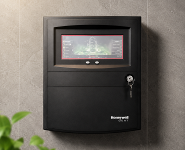 Wall-mounted black security panel with a small touchscreen display and a key in the lock; Honeywell Gent branding at the bottom.
